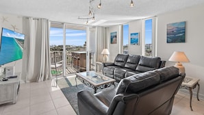A spacious living room with a large flat-screen TV, leather sofas, a glass coffee table, and a balcony with a view of the ocean. - Dolphin Point #406a (Destin)