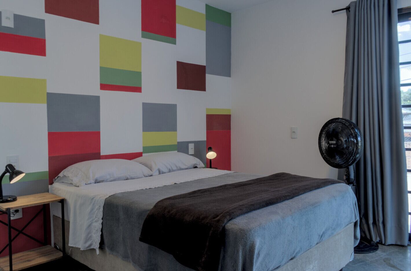 A bedroom with a bed, a bedside table with a lamp, a standing fan, and a window with curtains.