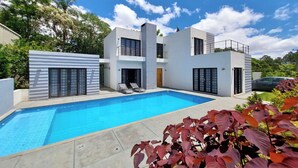 A modern two-story house with a swimming pool, a car parked nearby, and a garden with red foliage.