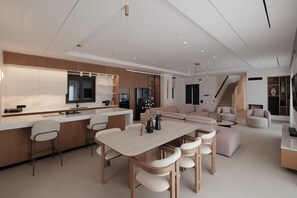 A modern kitchen with a central island, a dining area with a wooden table and chairs, and a living area with a sofa and armchairs. - Azura Exclusive Villa (Acharavi)