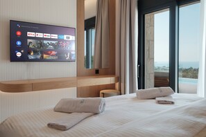 Ein Hotelzimmer mit einem großen Flachbildfernseher, der Streaming-Dienste anzeigt, einem Holz-Kopfteil, einem Bett mit ordentlich gefalteten Handtüchern und einem Balkon mit Blick.