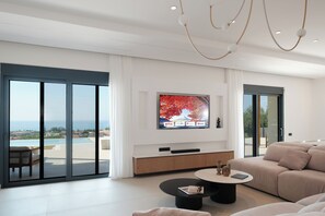 A modern living room with a large TV, a sofa, a coffee table, and a view of the sea through large sliding doors. - Azura Exclusive Villa (Acharavi)