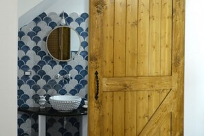 Een badkamer met een ronde spiegel, een witte wastafel en een wand betegeld met blauwe en grijze tegels.