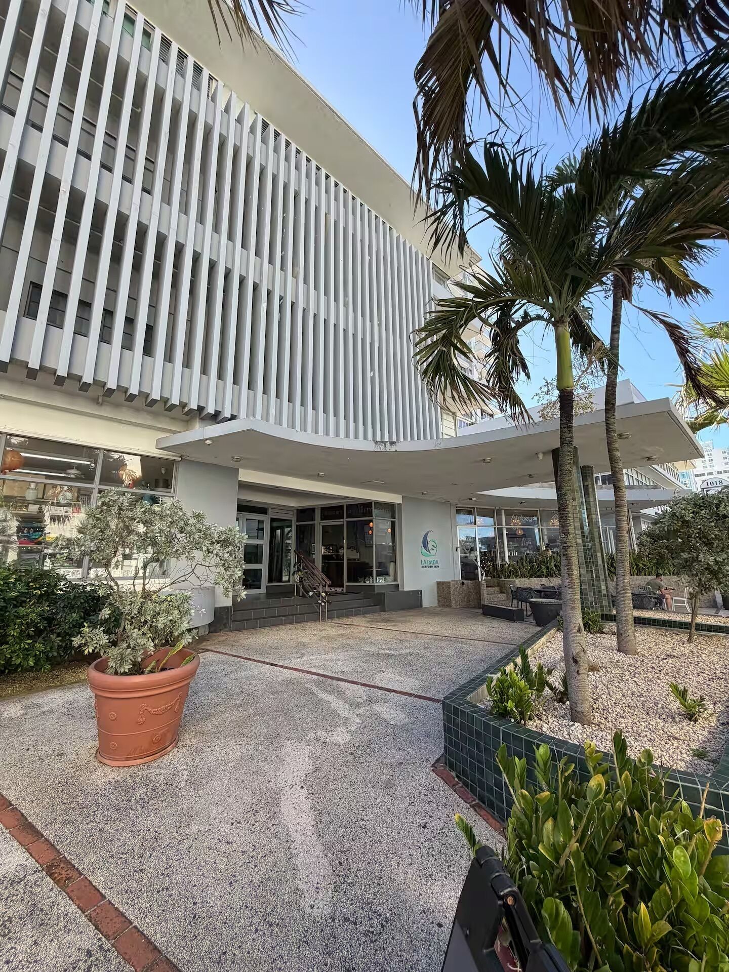 A modern building with a flat roof, large windows, and a paved entrance area with potted plants and a seating area.