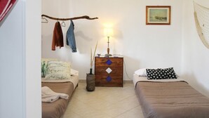 Une petite chambre bien rangée, avec deux lits simples, une commode en bois surmontée d’une lampe et un tableau accroché au mur.