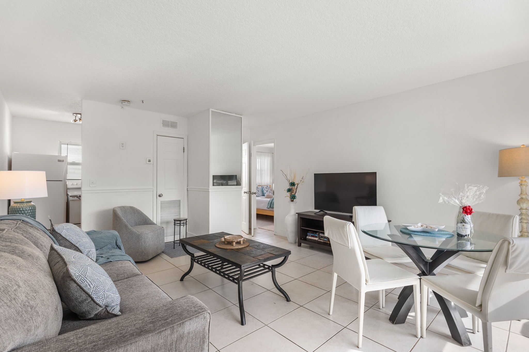 Un salon moderne avec un canapé, une table basse en verre, une table à manger avec des chaises, un téléviseur à écran plat et, au fond, un coin cuisine.