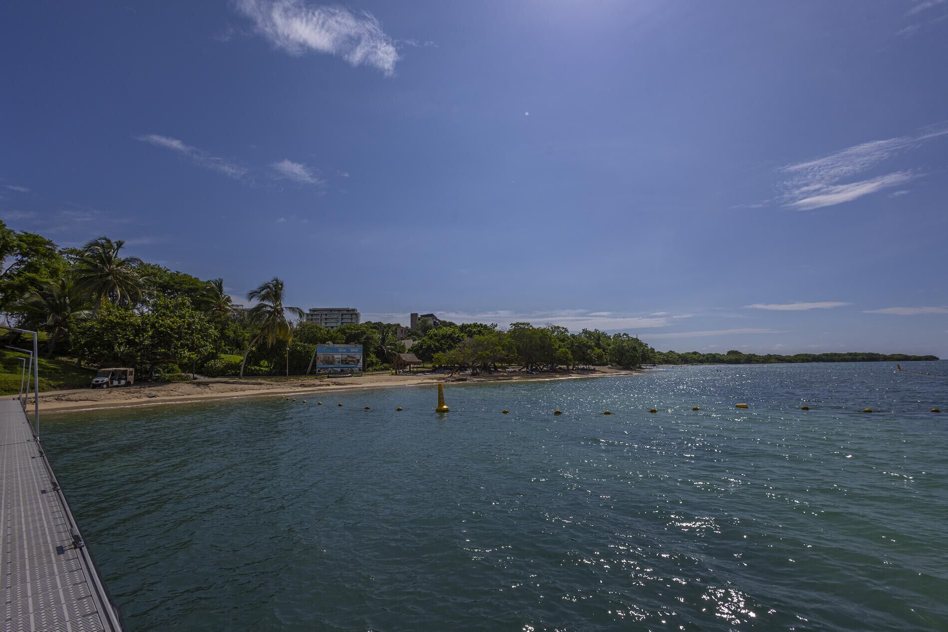 A coastal scene with a pier extending into calm waters, a sandy beach, and dense greenery including palm trees.