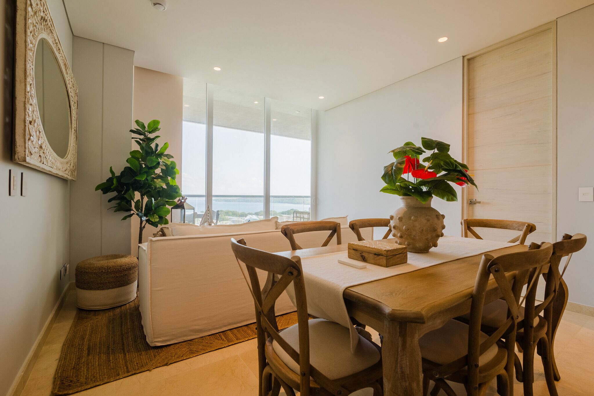 A dining area with a wooden table, six chairs, a vase with flowers, and a large window offering a view.