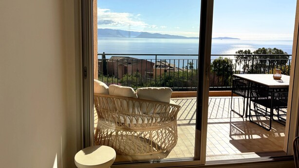 Un balcón con una silla de mimbre y una mesita pequeña, con vistas a un paisaje marino y montañas lejanas.
