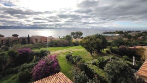 Un paisaje costero con vista al mar, un jardín con diversas plantas y edificios con tejados de terracota.