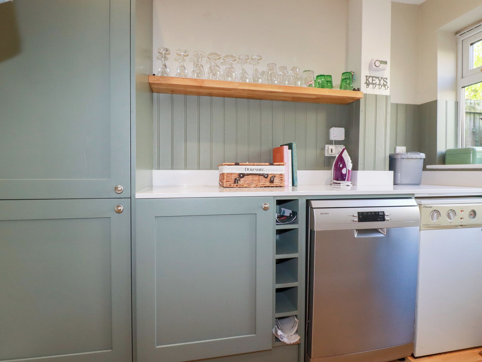 Una cocina con lavavajillas, plancha y una estantería con cristalería y libros.