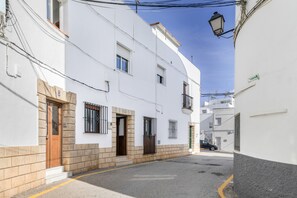 Uma rua estreita com edifícios brancos, uma porta de madeira, janelas com grades e um carro estacionado.
