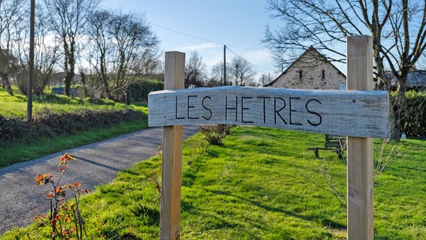 A wooden sign with 'LES HETRES' written on it, a paved path, and a grassy area with trees and a building in the background.