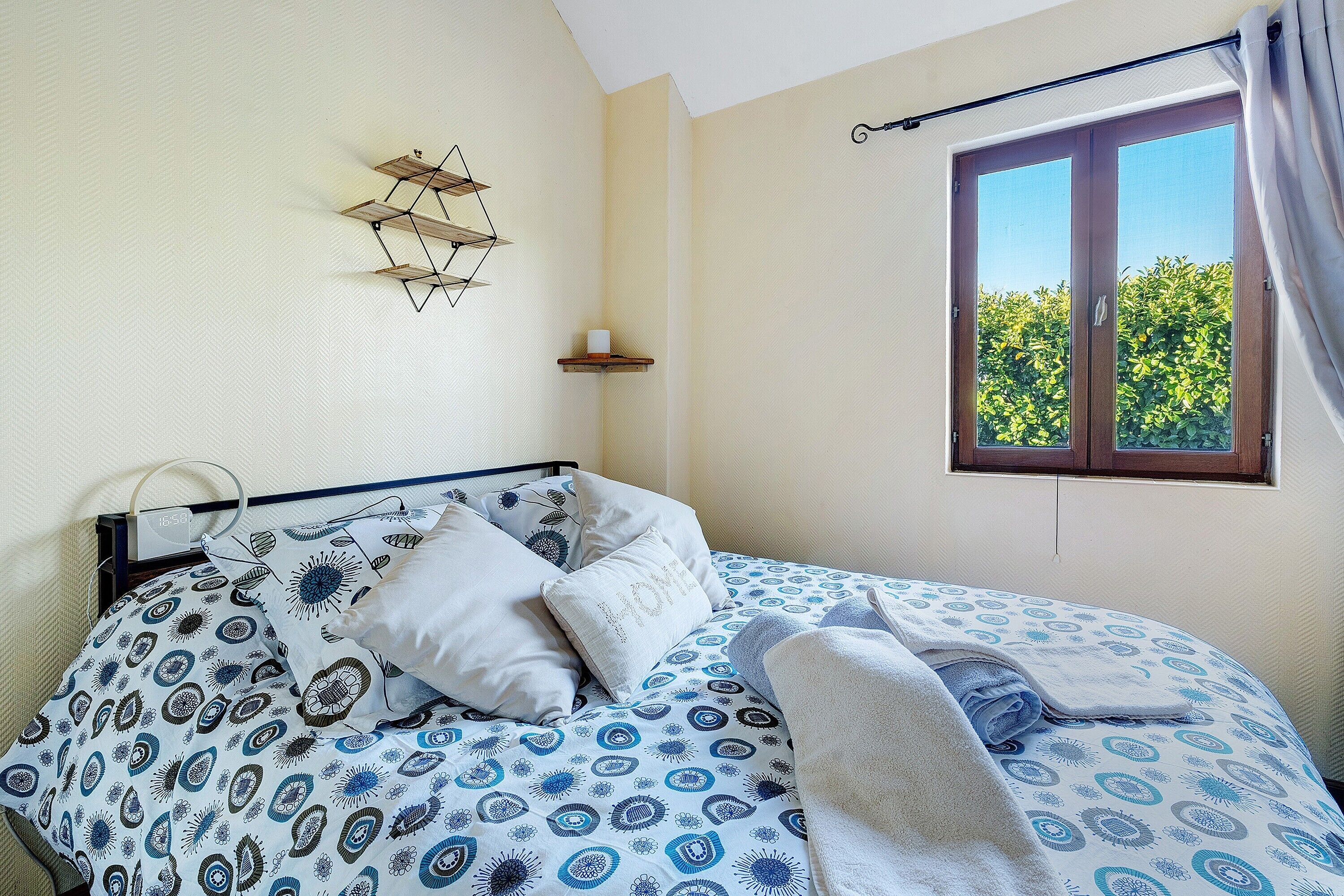 Un dormitorio con cama, almohadas, una mesita de noche, una ventana con cortinas y un estante en la pared.