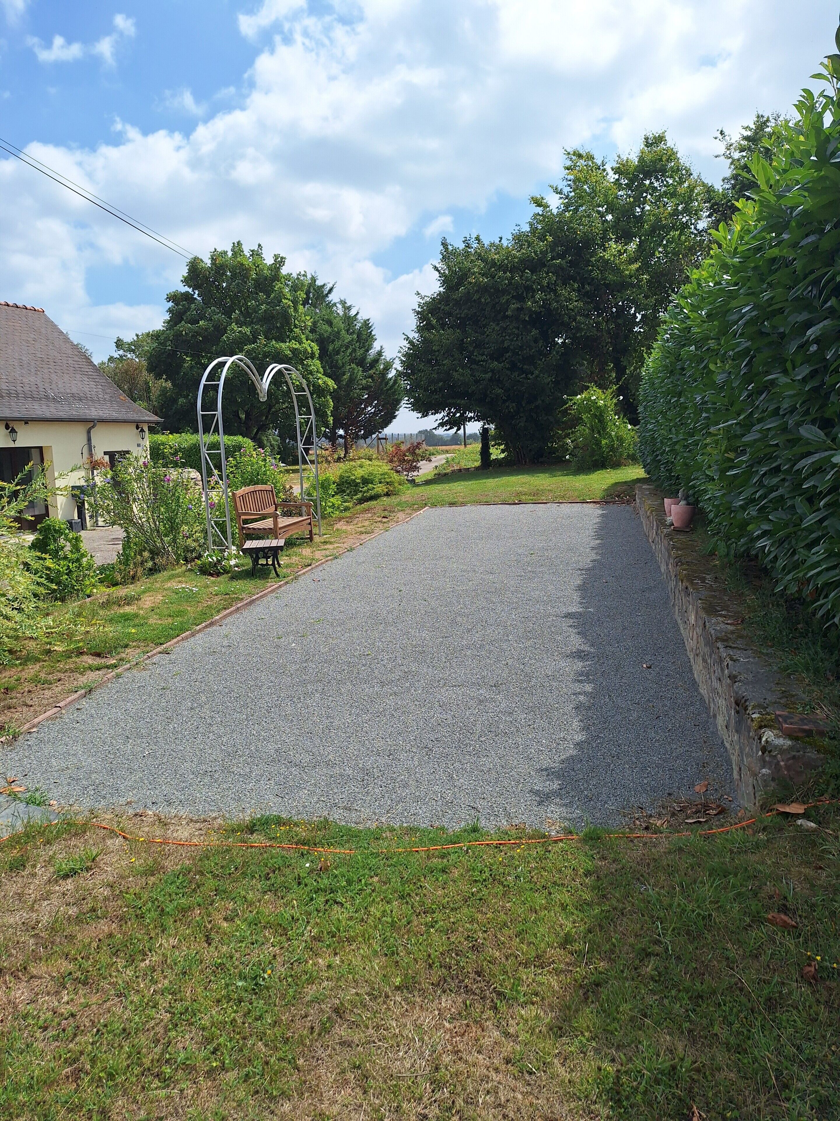 A paved driveway leading to a house with a garden, a wooden bench, and a metal archway.