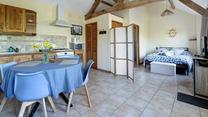 A kitchen with wooden cabinets, a dining table with chairs, a bed with a patterned bedspread, and a view of the outdoors.
