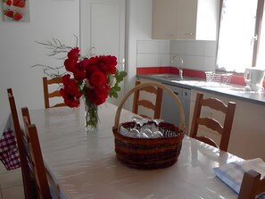Ett matbord med stolar, en vas med röda blommor, en korg med mat och en köksdel med en diskho och skåp.