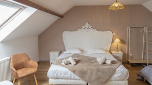 Un dormitorio con techo inclinado, una cama grande con ropa blanca, un edredón marrón, cabecera blanca, mesita de noche con lámpara, una silla de madera y un radiador.