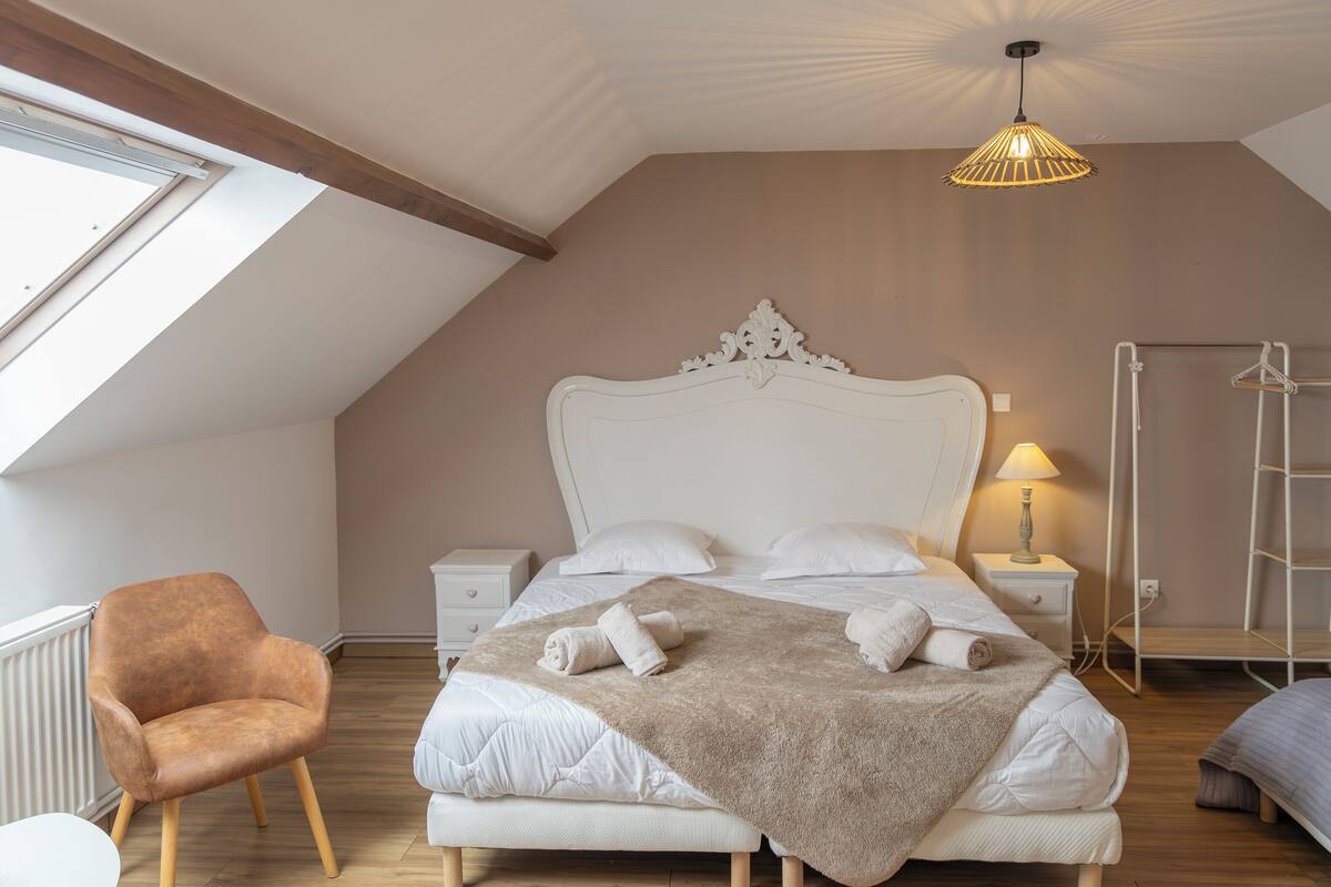 Un dormitorio con techo inclinado, una cama grande con ropa blanca, un edredón marrón, cabecera blanca, mesita de noche con lámpara, una silla de madera y un radiador.