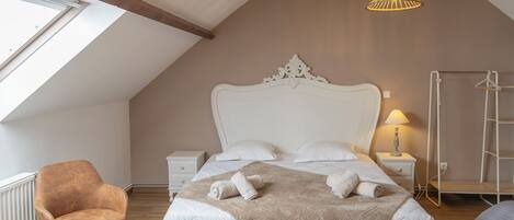 A bedroom with a sloped ceiling, a large bed with white bedding, a brown throw blanket, a white headboard, a bedside table with a lamp, a wooden chair, and a radiator.