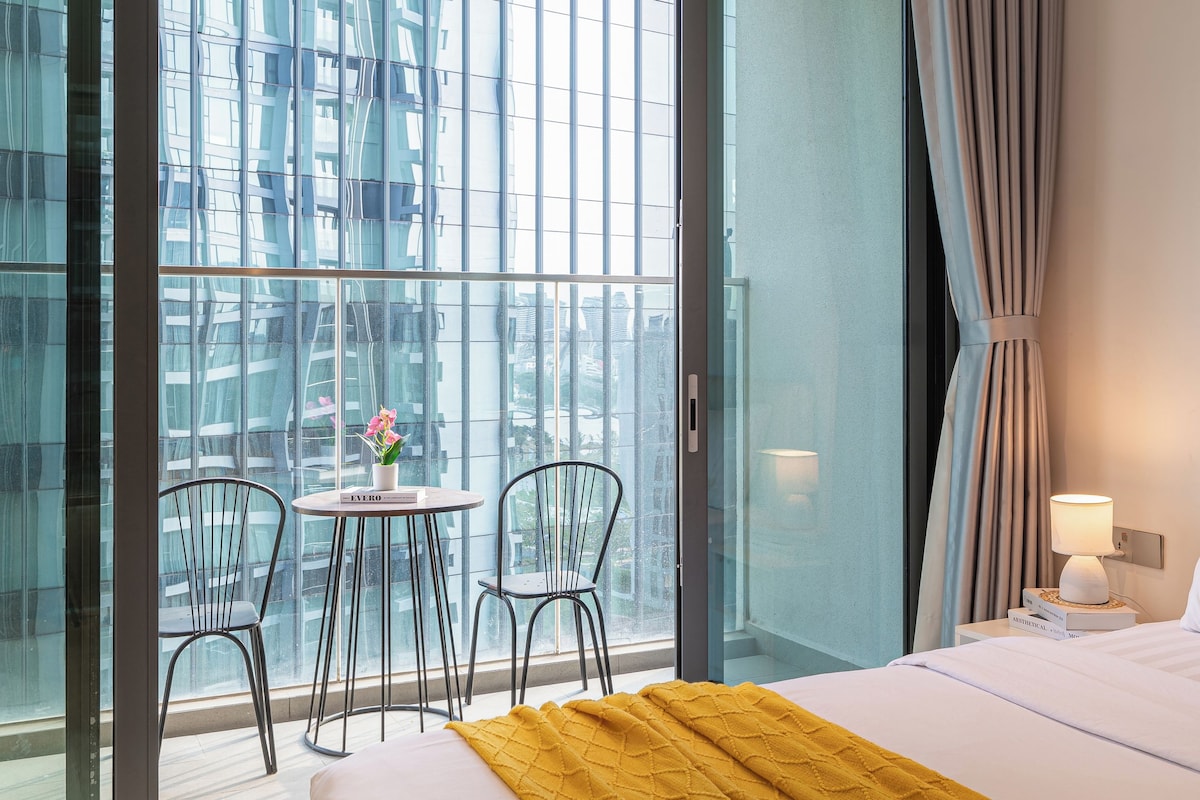 A hotel room with a bed, a small table with chairs, a balcony with a view of buildings, and a window with curtains.