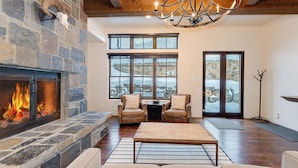 A cozy living room with a stone fireplace, leather chairs, a coffee table, and large windows offering a view of a snowy landscape. - Luxury at Whitefish Mountain chalet w/ hot tub (Whitefish)