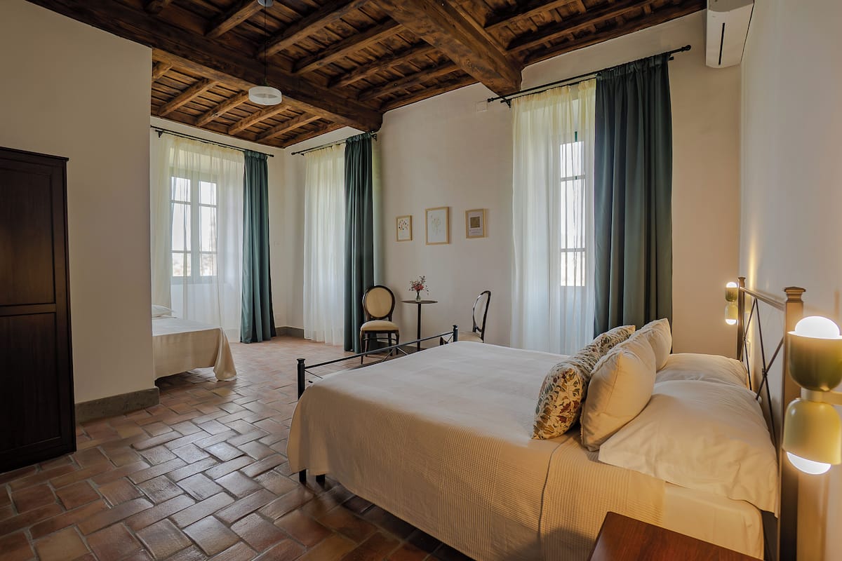 A bedroom with a large bed, wooden beams on the ceiling, a small table with chairs, and a window with curtains.