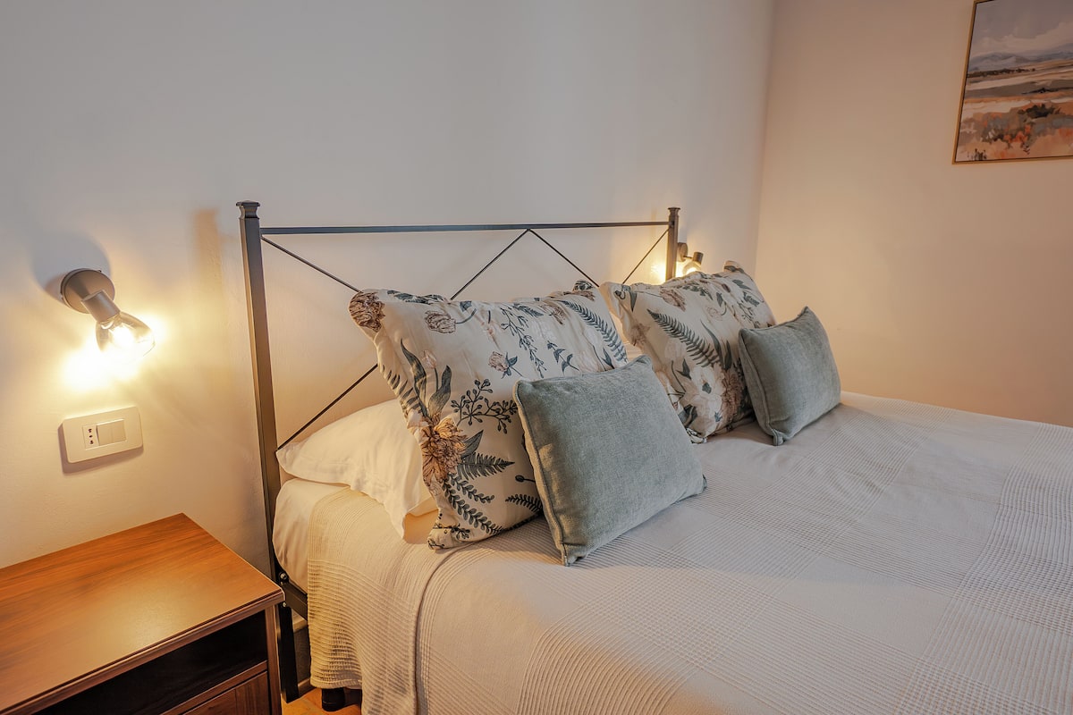 A neatly made bed with floral patterned pillows, a wooden nightstand, and a wall-mounted light fixture.