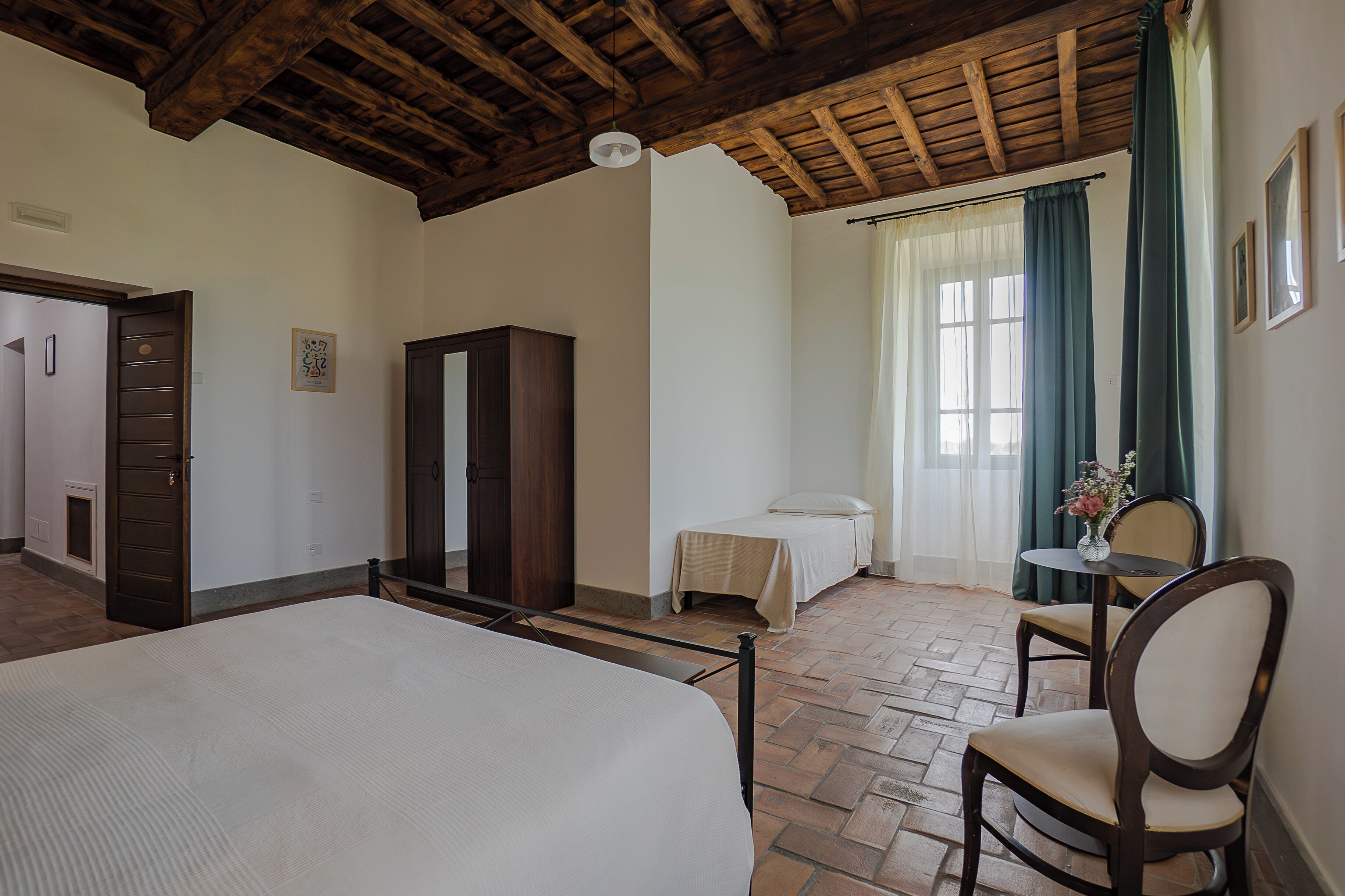 A bedroom with a bed, a wooden wardrobe, a small table with a vase, and a chair.