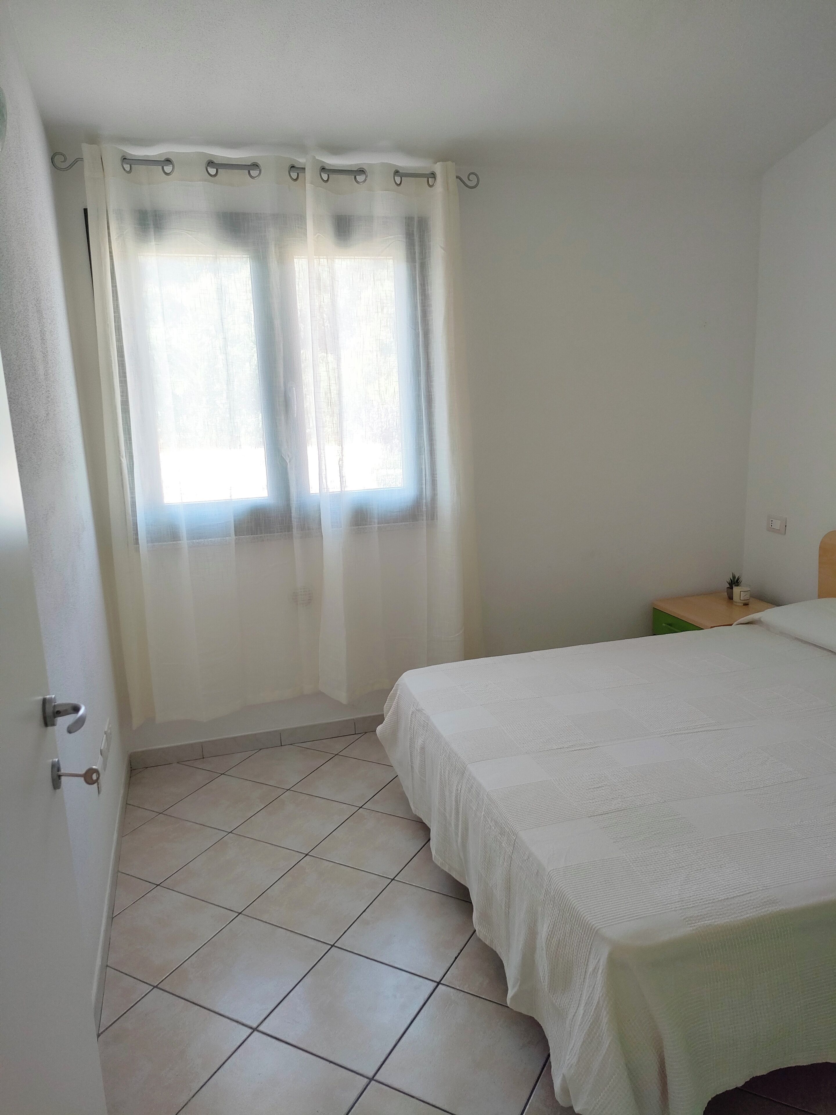 Une chambre à coucher avec un lit, une table de chevet, une commode verte et une fenêtre ornée de rideaux légers.