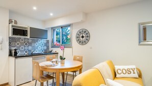 A compact kitchen with a microwave, a sink, and a window. There's a dining table with chairs and a yellow sofa with a 'COSY' pillow. A decorative clock is on the wall.