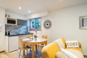 Eine kompakte Küche mit Mikrowelle, Spüle und Fenster. Ein Esstisch mit Stühlen und ein gelbes Sofa mit einem 'COSY'-Kissen. Eine dekorative Uhr befindet sich an der Wand.