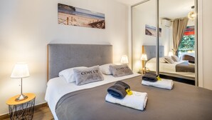 A hotel room with a bed, two pillows, a bedside lamp, a small table, a mirror, and a beach painting on the wall.
