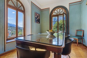 A dining area with a glass table, chairs, a bowl of fruit, and a view of mountains through large windows.