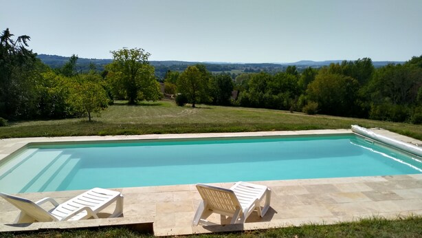 Piscina rectangular con dos sillas reclinables blancas sobre un deck de piedra, rodeada de césped y árboles, con vistas a colinas distantes.