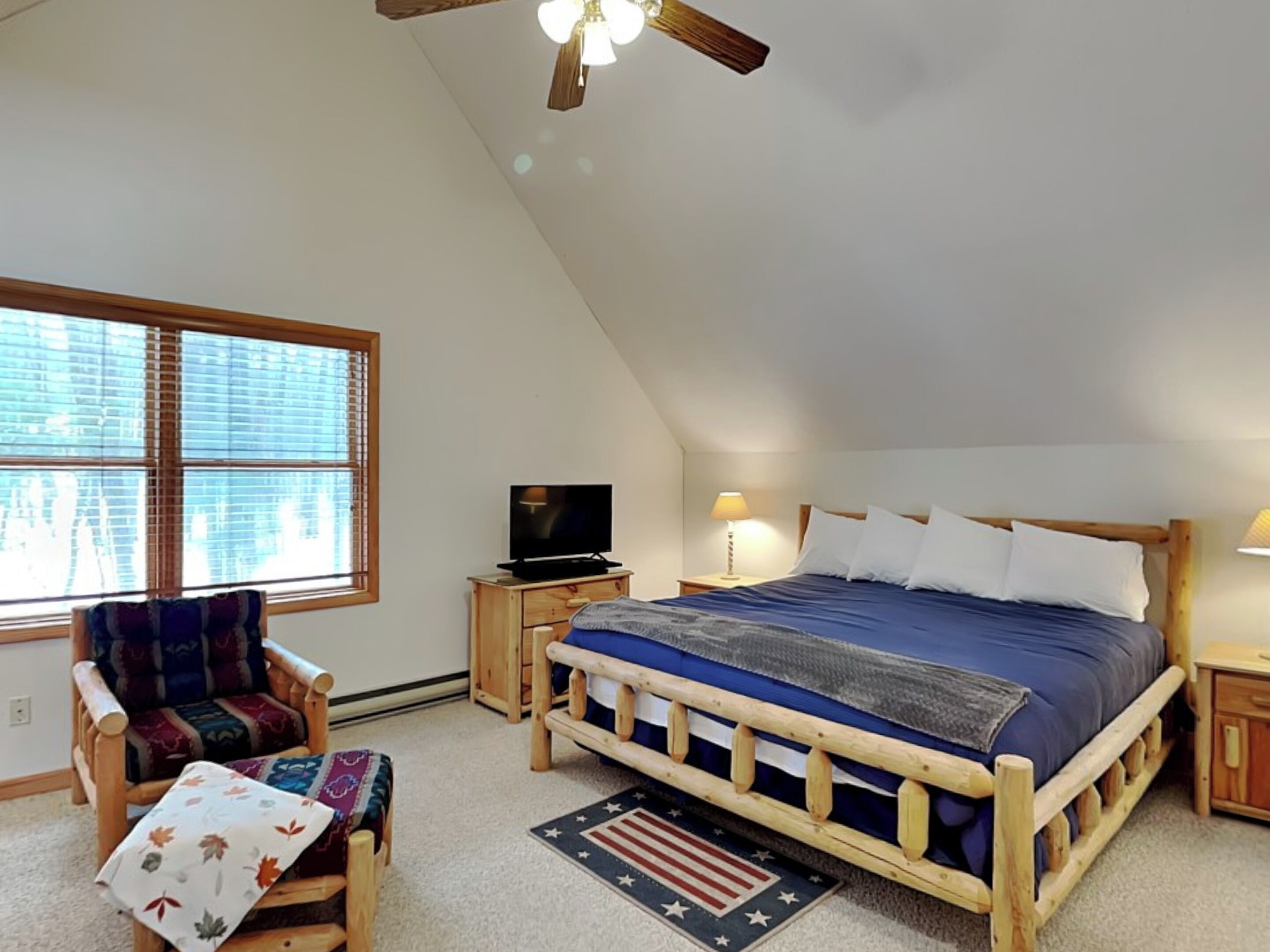 A bedroom with a sloped ceiling, a bed with a wooden frame, a television on a wooden nightstand, a chair with a patterned cushion, and a window with blinds.