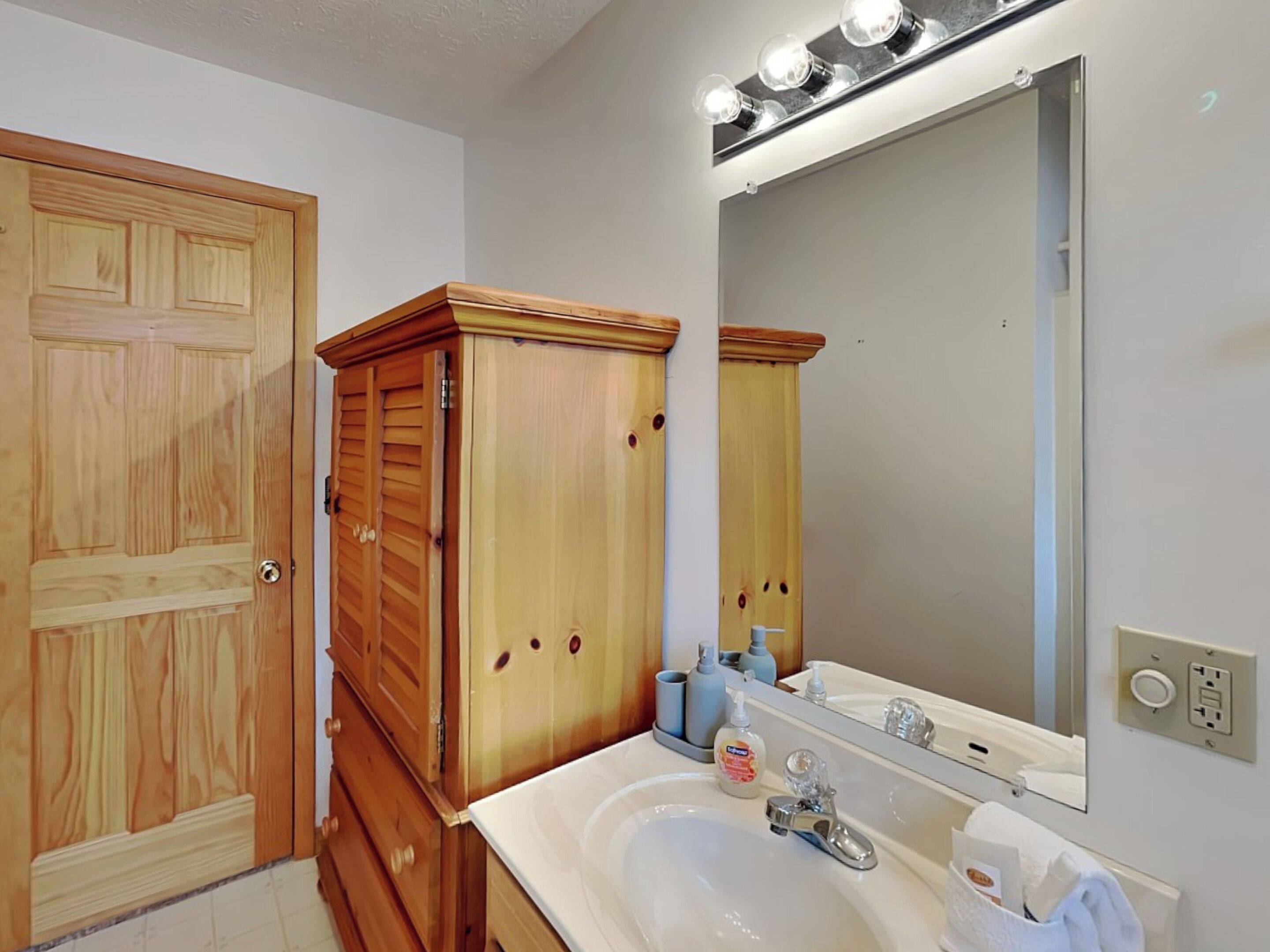 A bathroom with a wooden vanity, a sink, a mirror with lights, and toiletries.