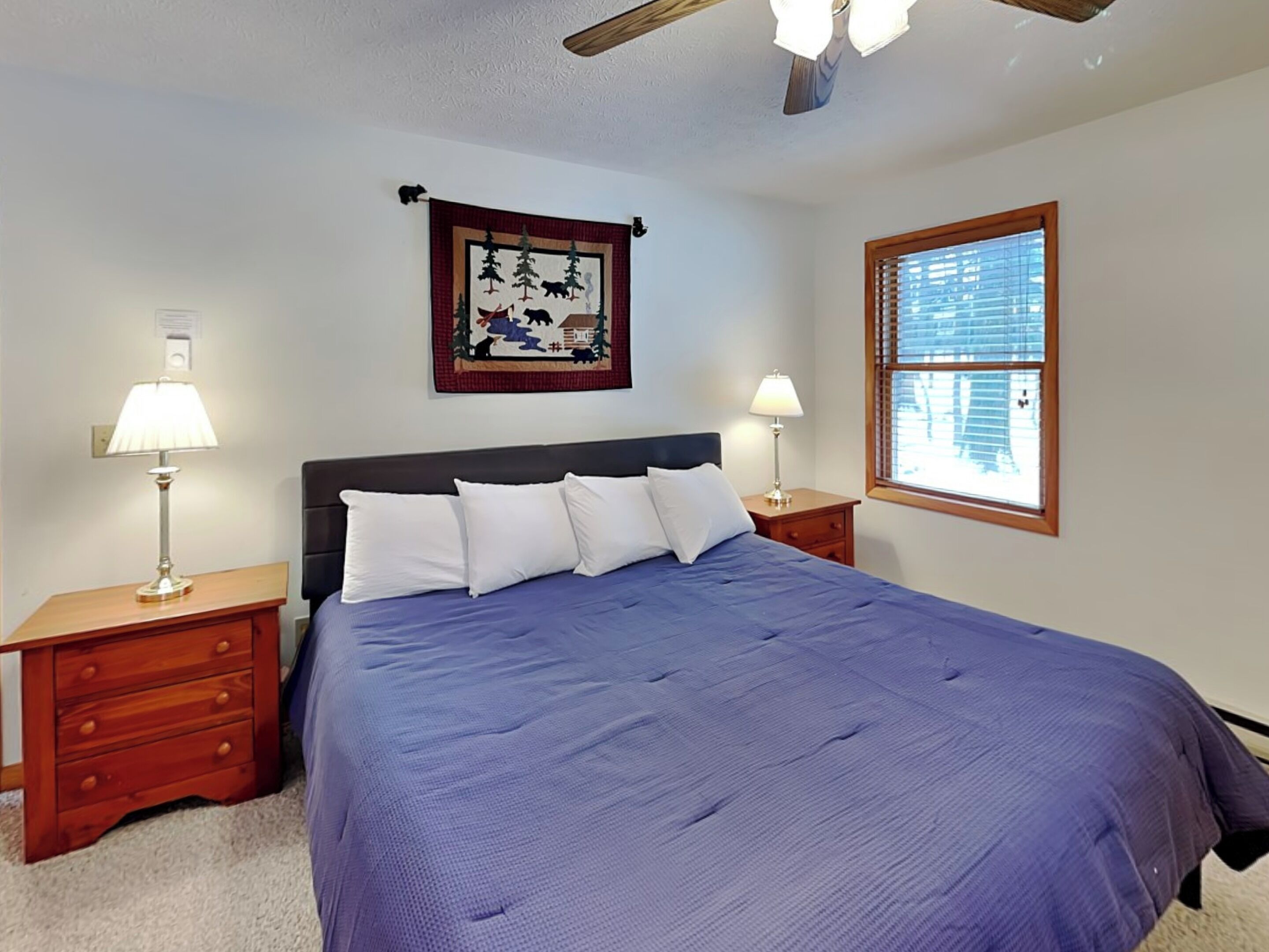 A bedroom with a large bed, two bedside tables with lamps, a ceiling fan, a window with blinds, and a wall hanging.