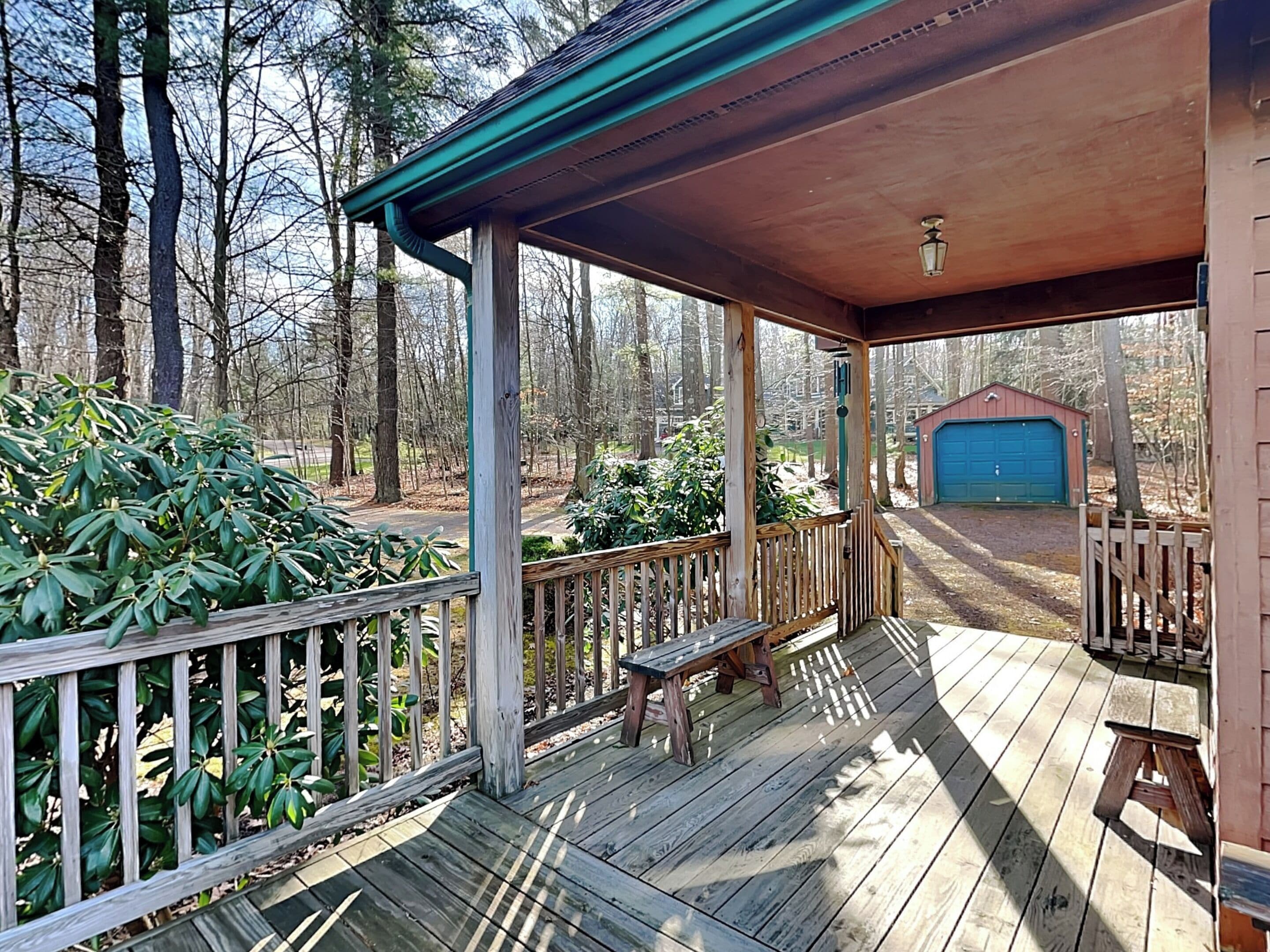 A wooden deck with a bench and a covered area, surrounded by trees and a garage.