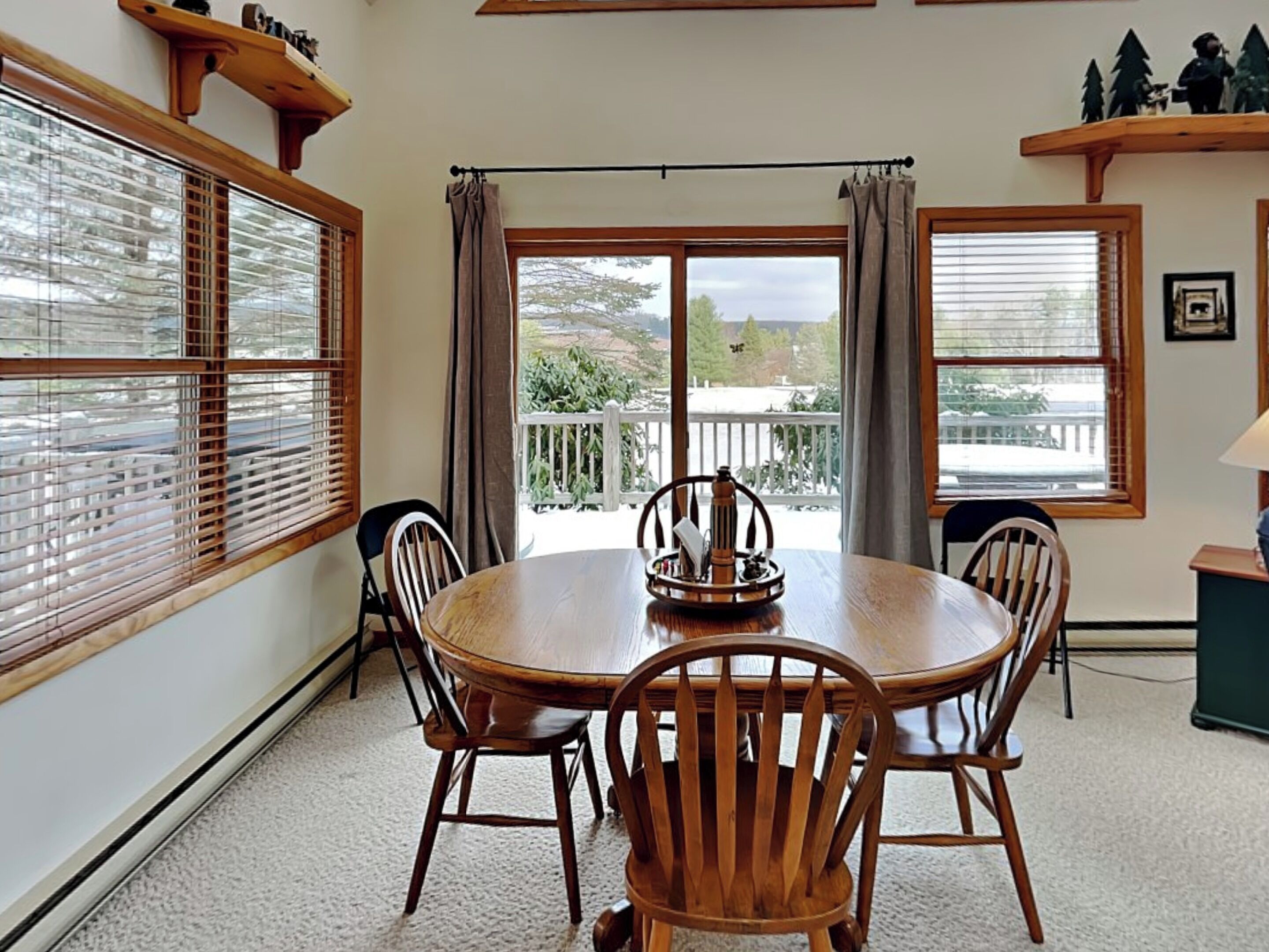 A dining area with a round table and four wooden chairs, large windows with blinds, and a view of a balcony with a railing.