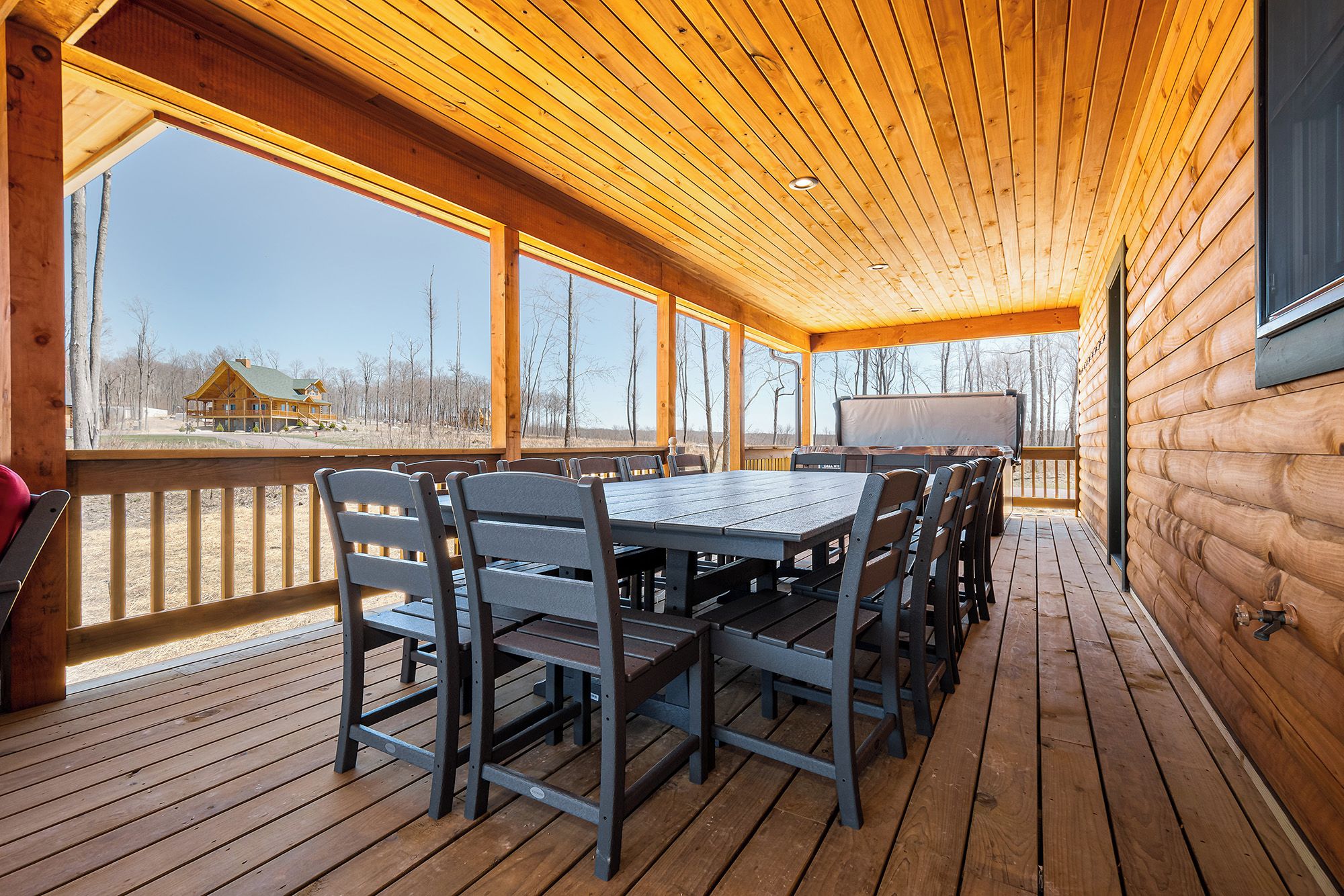 A covered outdoor deck with a wooden table and chairs, large windows, and a view of a cabin in the distance.