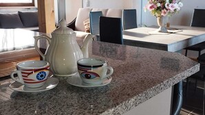A kitchen area with a granite countertop, a white kettle, two cups with decorative lids, and a vase with flowers on the dining table. - Vacation apartment in the Franconian Forest (Nordhalben)