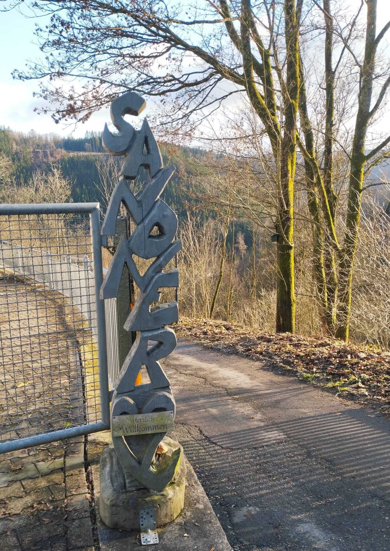 A metal sign with the inscription "WILLKOMMEN" at a gate, surrounded by trees and a fence.