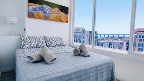 A bedroom with a bed, pillows, and a view of buildings through large windows. - Skyline Tower (Torre del Mar)