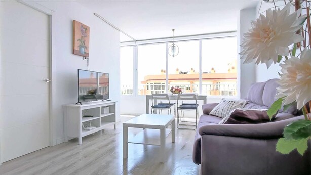 A modern living room with a sofa, a TV on a white stand, a dining table with chairs, and large windows offering a city view. - Skyline Tower (Torre del Mar)