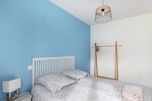 A bedroom with a bed, a bedside table with a lamp, a hanging light fixture, and a wooden rack.