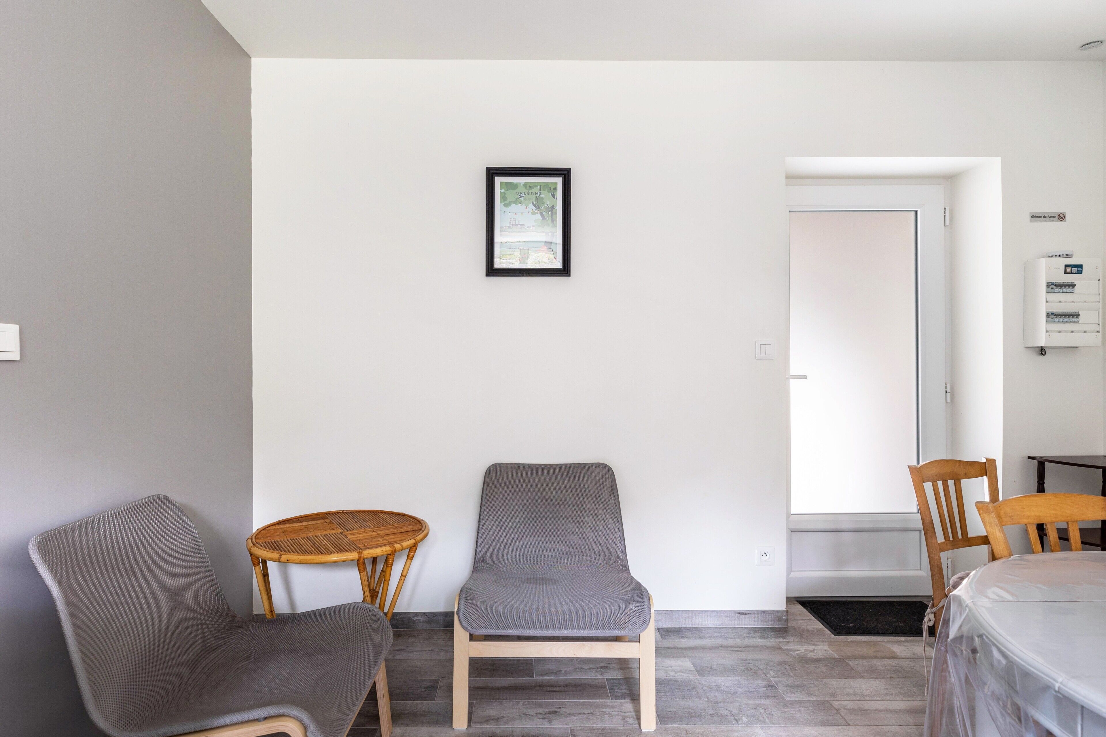 A room with a grey cushioned chair, a wicker chair, and a wooden chair. There is a framed picture on the wall, a door, and a white air conditioning unit.