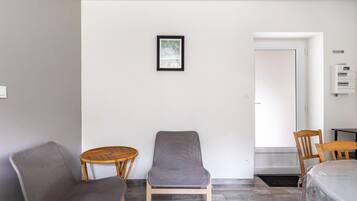 A room with a grey cushioned chair, a wicker chair, and a wooden chair. There is a framed picture on the wall, a door, and a white air conditioning unit.
