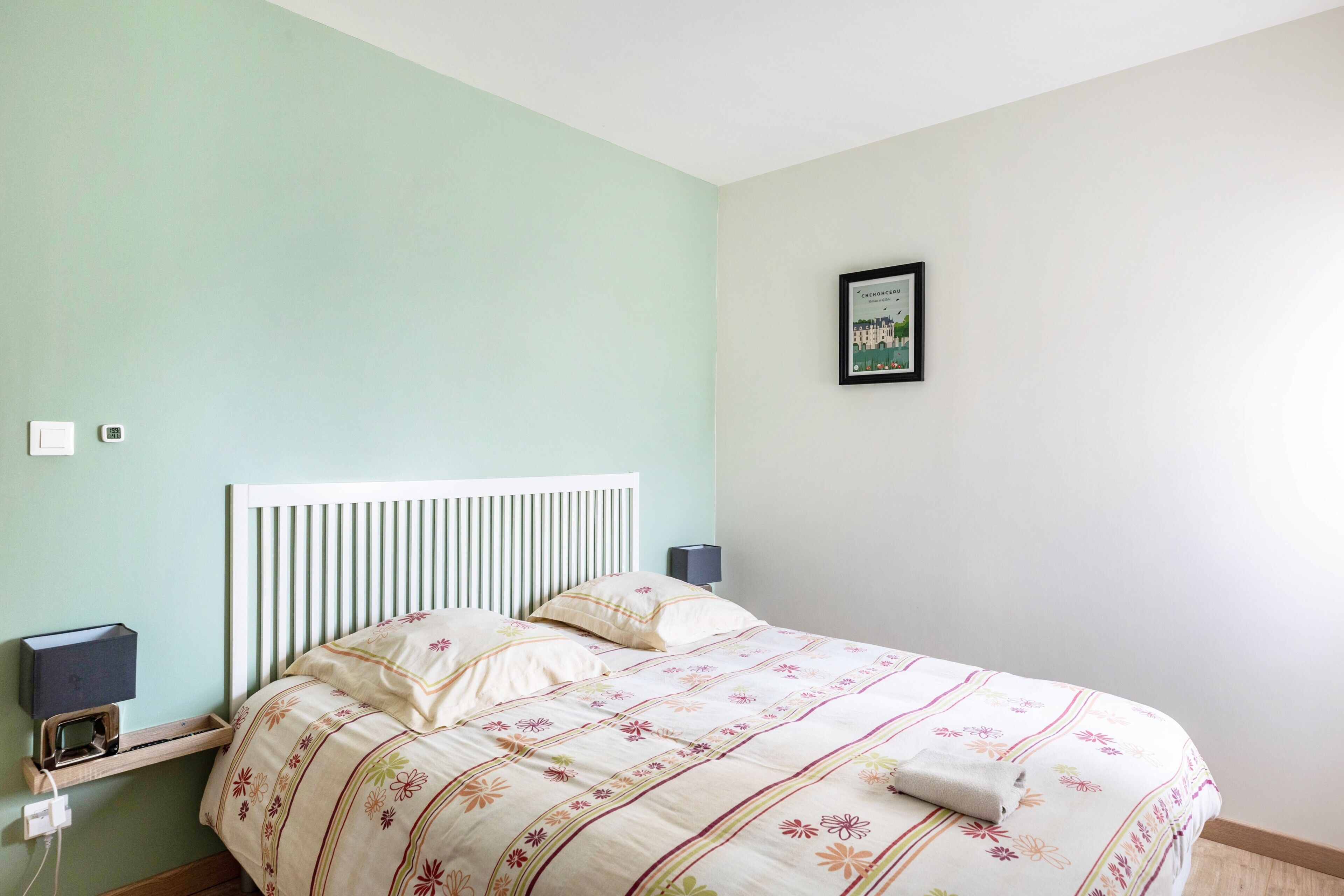 Una cama con colcha estampada, una mesita de noche con lámpara y un cuadro enmarcado en la pared.