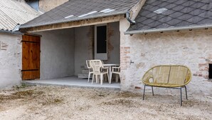 A rustic building with a wooden door, a small table with chairs, and a wicker chair outside.